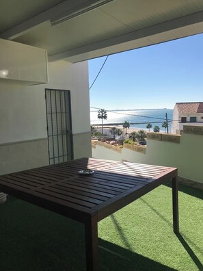 Un balcon avec une table en bois et un bol en métal, donnant sur une plage bordée de palmiers et offrant une vue sur un ciel dégagé.