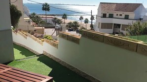 Uma varanda com grama sintética, uma mesa de madeira e vista para o mar e edifícios.
