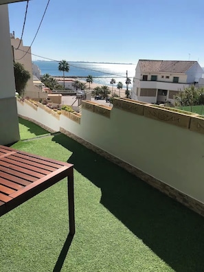 A balcony with artificial grass, a wooden table, and a view of the sea and buildings. - Apartment with Ocean Views in Fuentebravía Beach (EL PUERTO DE SANTA MARIA)