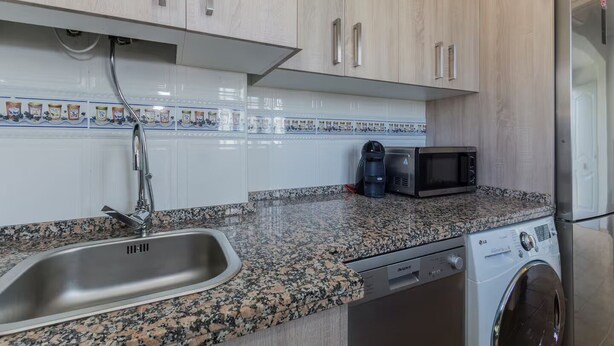 Uma cozinha com bancada de granito, pia de aço inoxidável, micro-ondas, cafeteira e máquina de lavar.