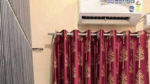 Ein Zimmer mit einem Bett, einem blauen Kopfteil, einem weißen Kissen, einem roten Vorhang mit Blätermotiv und einer Klimaanlage an der Wand.