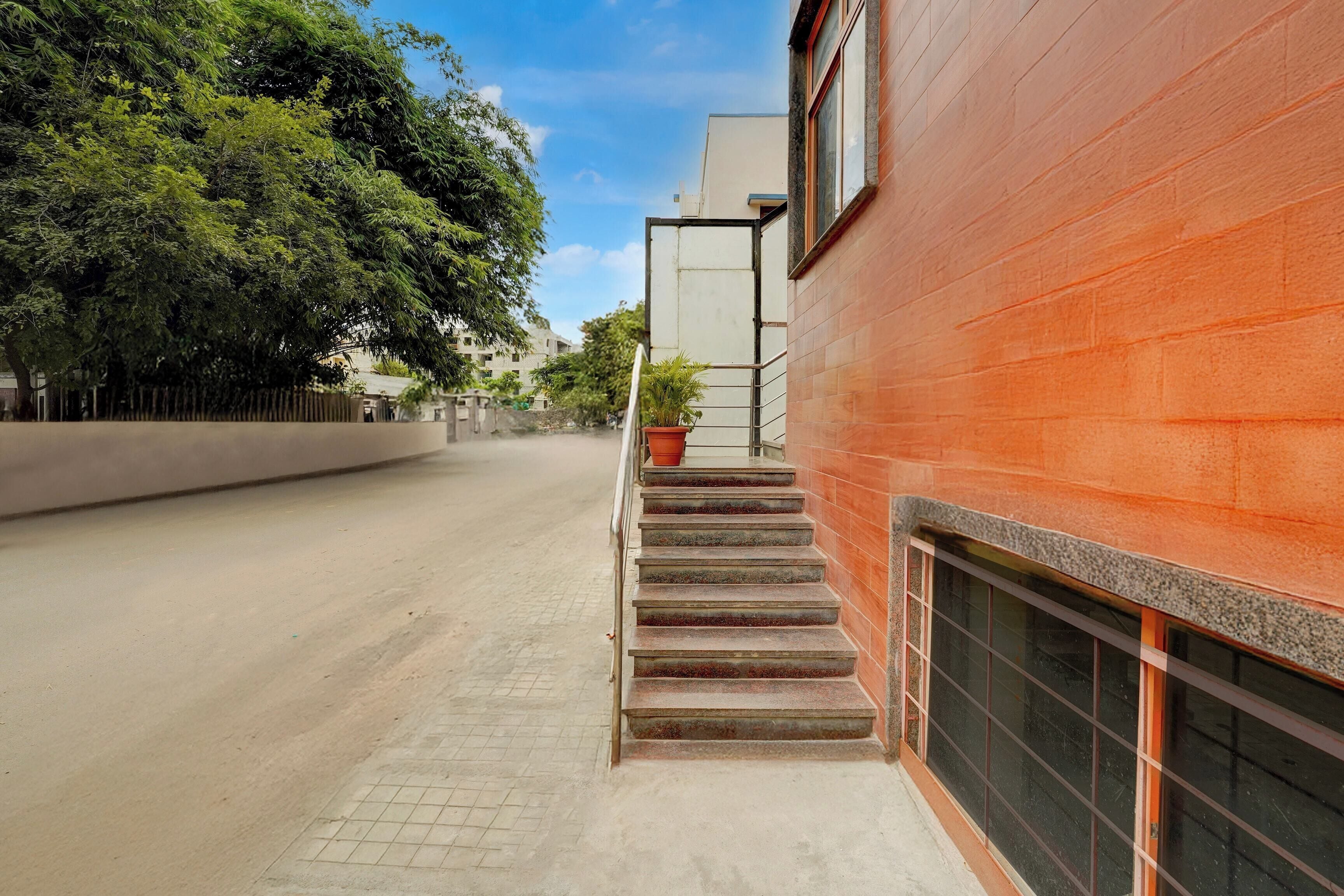 A narrow urban street with a red building on the right, a staircase leading to a door, and a tree-lined sidewalk.