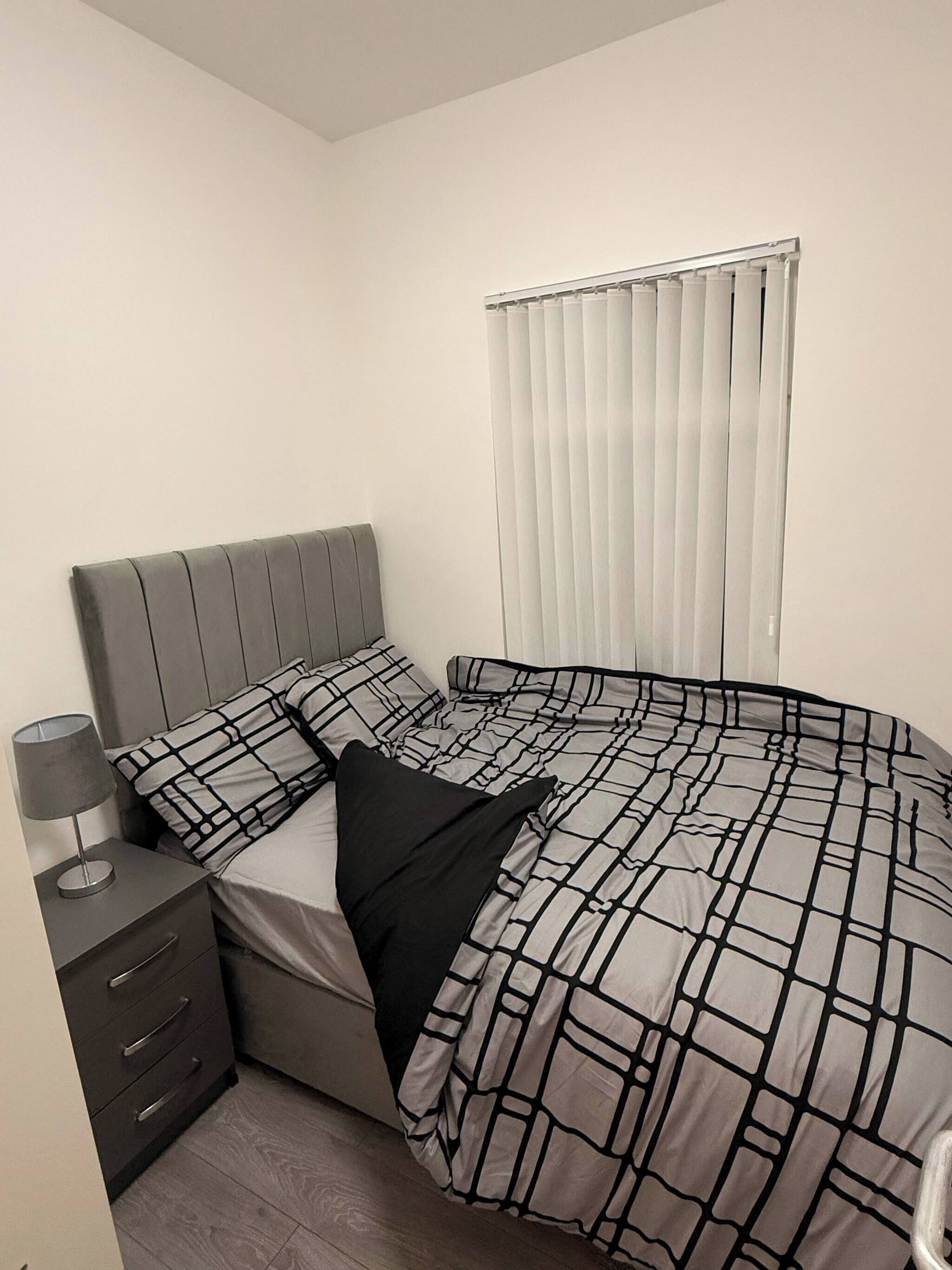 A bedroom with a bed, a nightstand, a lamp, a window with blinds, and a wooden floor.