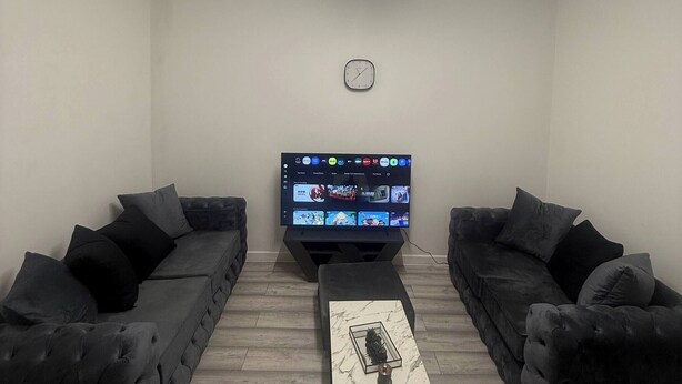 Una sala de estar con dos sofás oscuros, un televisor de pantalla plana, una mesa de centro de mármol y un reloj en la pared.