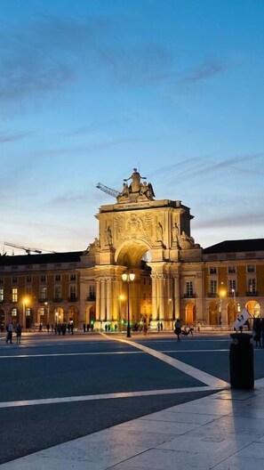 Ein großer, historischer Bogen mit aufwendigen Schnitzereien und einer Statue, umgeben von einem Platz mit einigen Personen und einem Mülleimer.