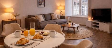 Un salon moderne avec une table à manger ronde, des canapés, un téléviseur et une fenêtre donnant sur des immeubles.