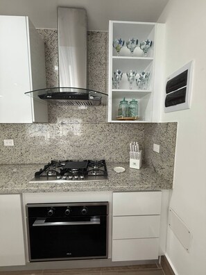 A kitchen with a gas stove, oven, white cabinets, a stainless steel range hood, and a shelf with glassware. - Sorelipeaceful Retreat at Green One Playa Dorada (Puerto Plata)