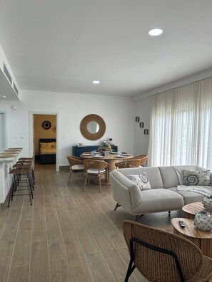 A modern living room with a sofa, dining table, and chairs. There is a kitchen area with a counter and stools. - Sorelipeaceful Retreat at Green One Playa Dorada (Puerto Plata)