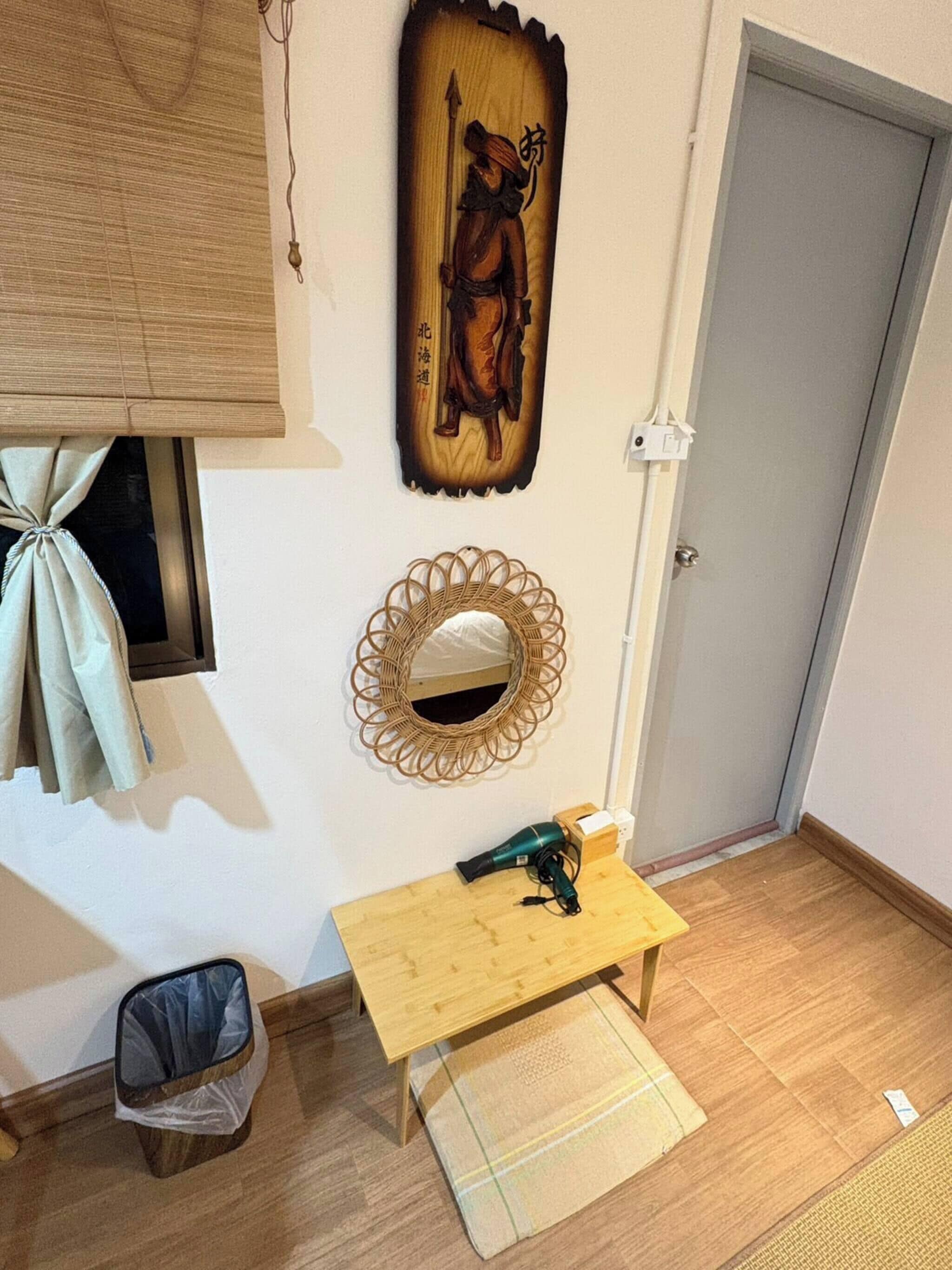 A room with a wooden floor, a small wooden table with a hairdryer, a woven mirror, and a wall-mounted artwork.