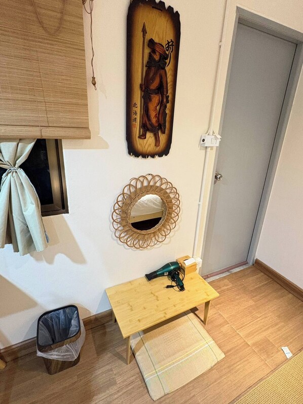 Una habitación con piso de madera, una pequeña mesa de madera con un secador de pelo, un espejo tejido y una obra de arte colgada en la pared.