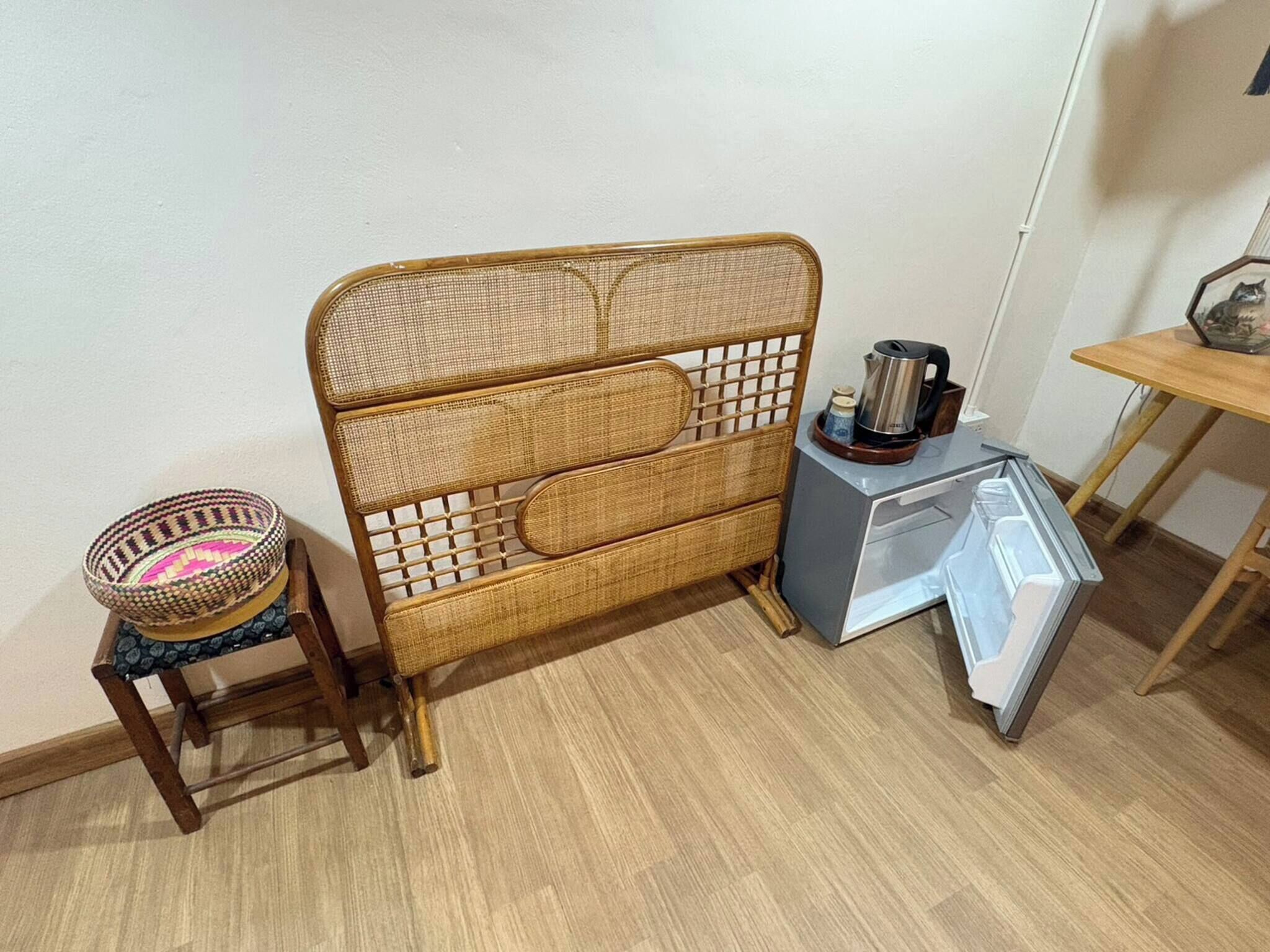 A room with a wicker headboard, a small wooden stool with a woven basket, a gray refrigerator, and a wooden table with a kettle and cups.