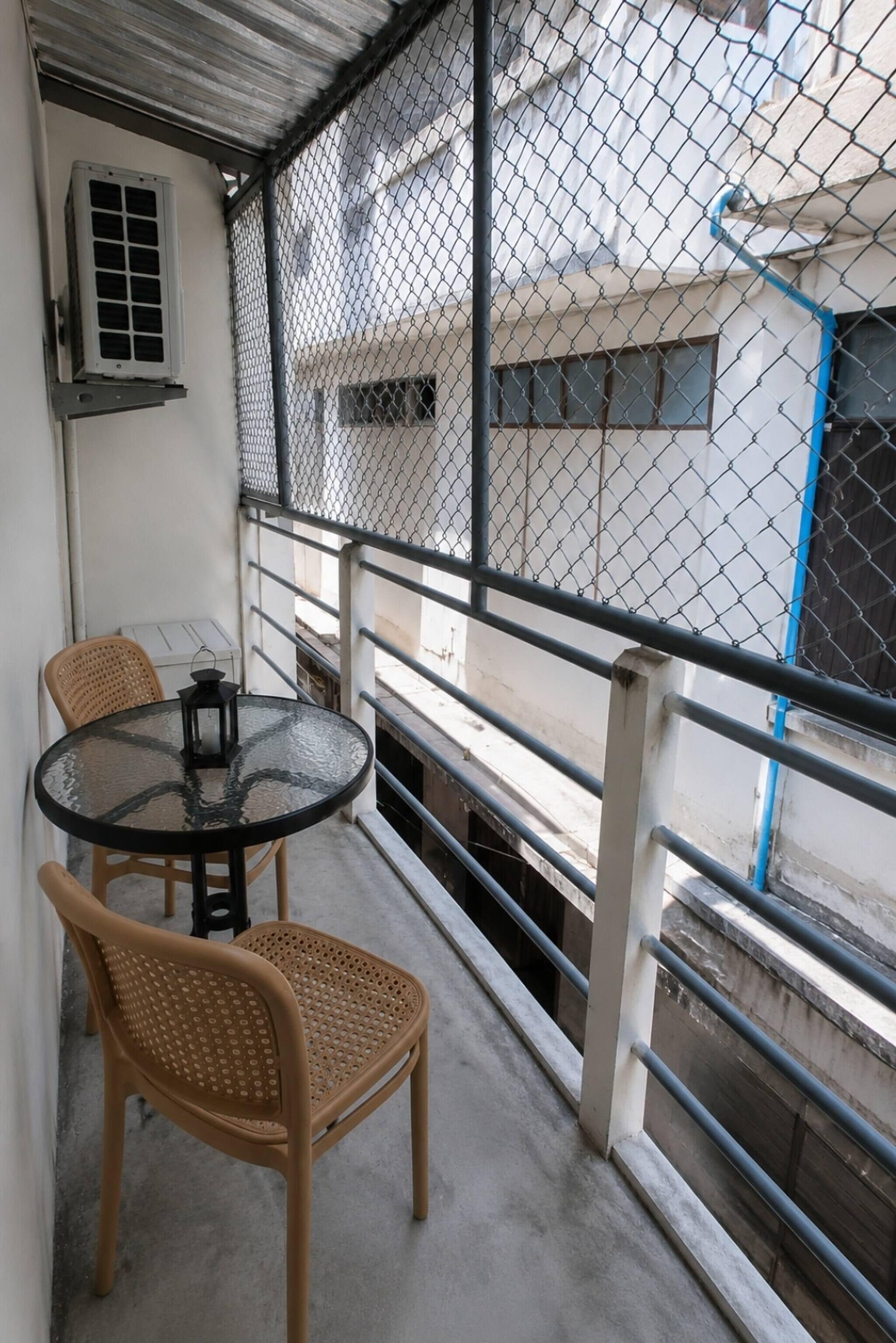 Een balkon met een ronde tafel en twee rieten stoelen, een hek van gaas en een gebouw op de achtergrond.