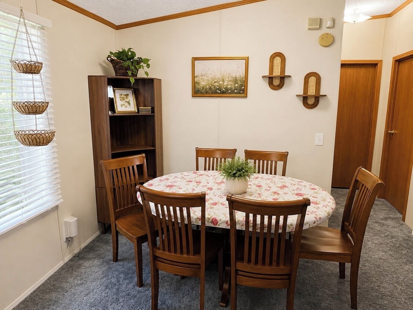 A dining area with a round table, wooden chairs, a floral tablecloth, a potted plant, a framed picture, and wall-mounted sconces.