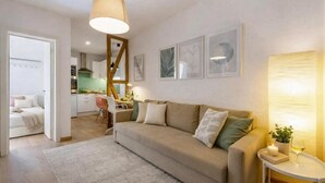 Ein modernes Wohnzimmer mit einer Couch, einem kleinen Tisch und einer Lampe. Im Hintergrund ist eine Küche zu sehen.