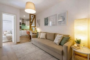 Ein modernes Wohnzimmer mit einer Couch, einem kleinen Tisch und einer Lampe. Im Hintergrund ist eine Küche zu sehen.