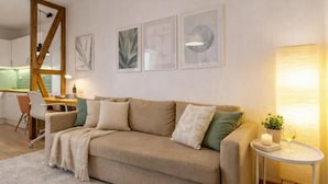 Ein modernes Wohnzimmer mit einem beigefarbenen Sofa, einem kleinen runden Tisch mit Kerze und Glas, und einer Küche im Hintergrund.