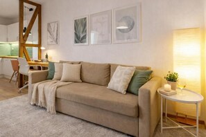 Ein modernes Wohnzimmer mit einem beigefarbenen Sofa, einem kleinen runden Tisch mit Kerze und Glas, und einer Küche im Hintergrund.