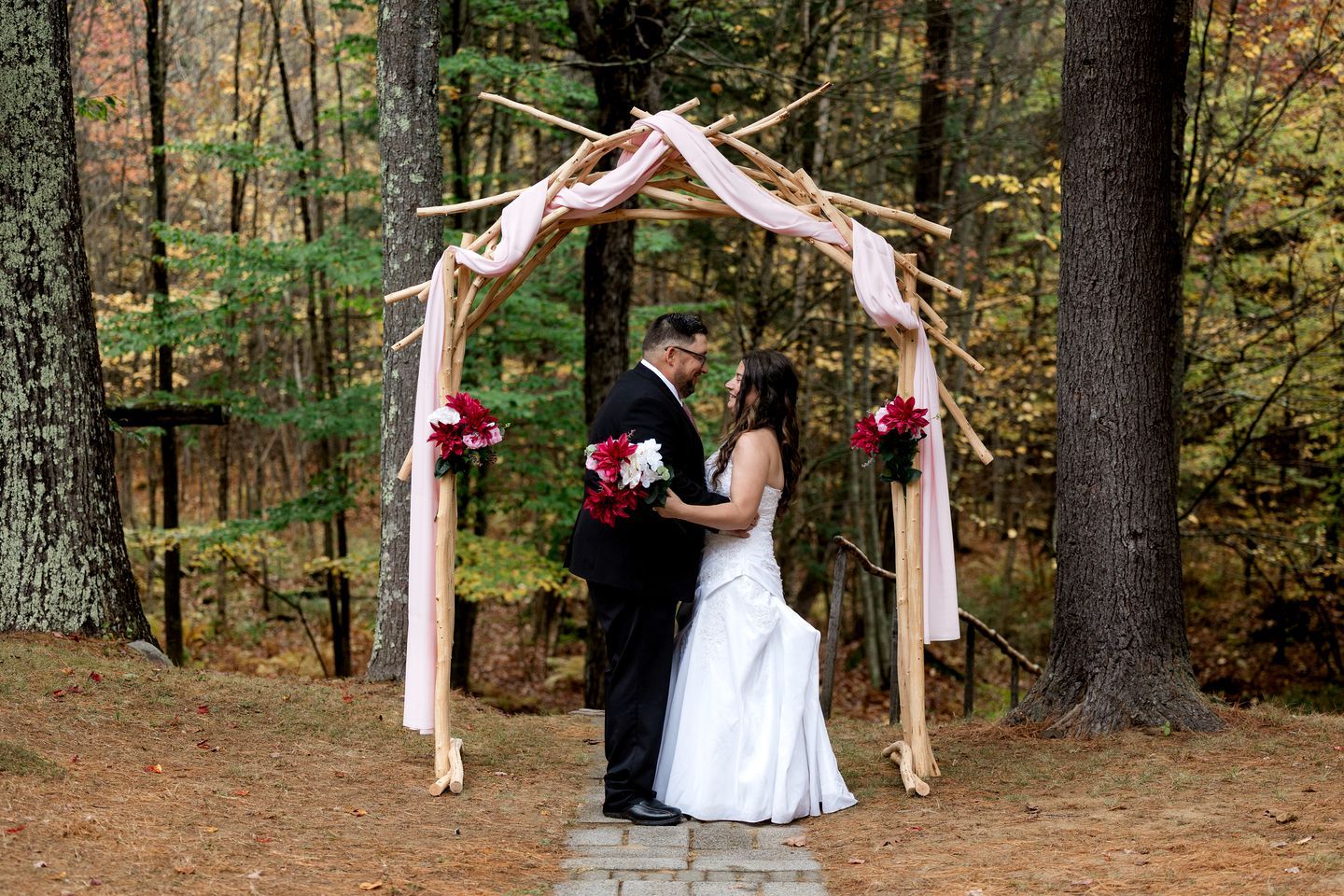A couple under a wooden archway with floral decorations in a forest setting.