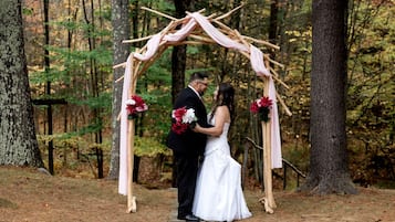 Un couple se tient sous une arche en bois, décorée de fleurs, dans un cadre forestier.