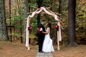 A couple under a wooden archway with floral decorations in a forest setting.