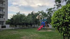 A playground with a red slide and a climbing structure, surrounded by trees and a building. - Spacious & Cozy 2BHK Retreat , Pet-Friendly , Near SIDCUL & Har Ki Pauri (Salempur Mahdood)