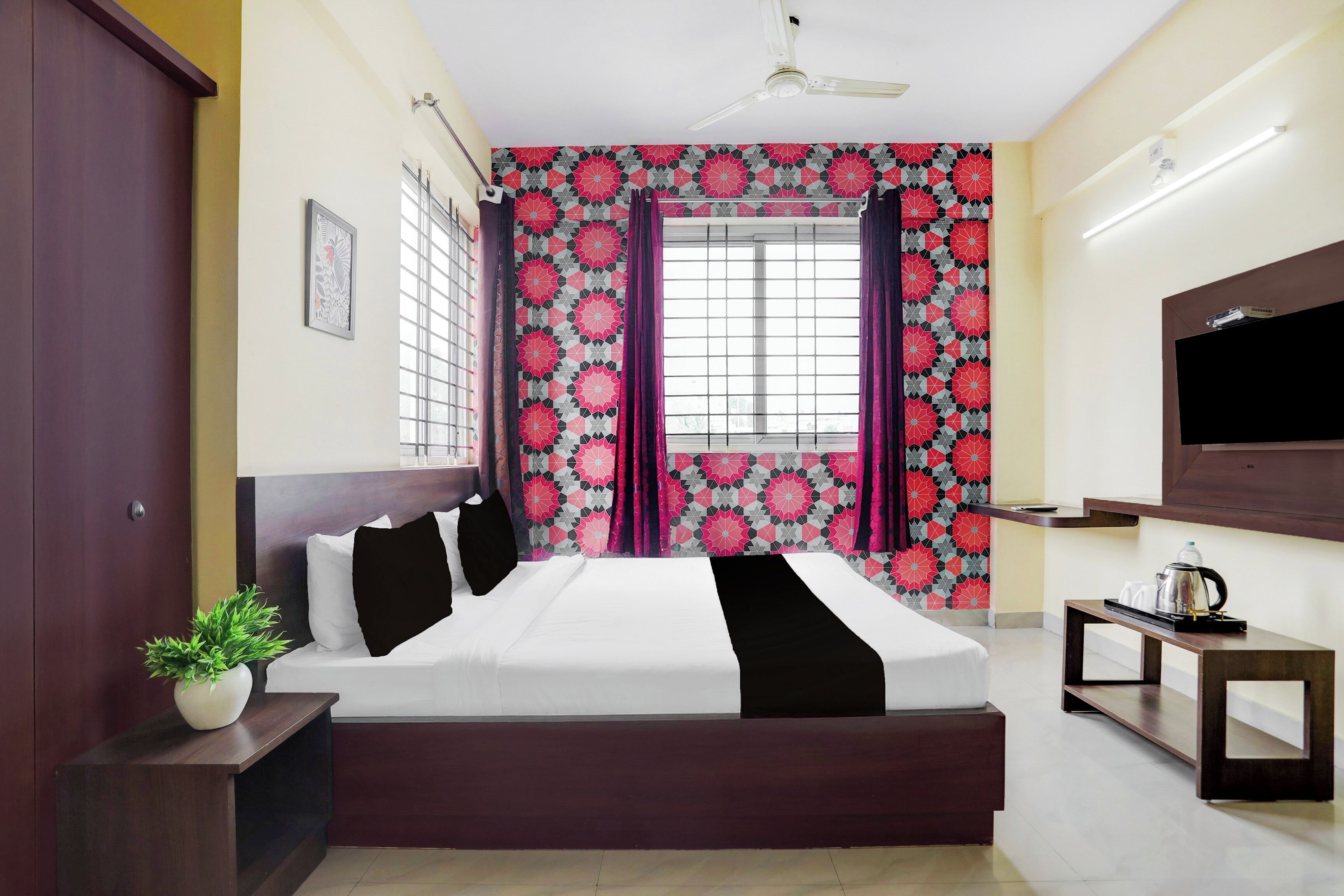 A hotel room with a bed, a TV, a bedside table with a kettle, and a window with curtains.