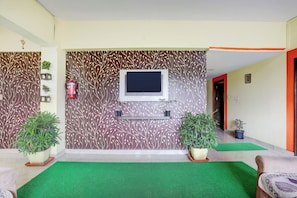 Un hall de hotel con una televisión de pantalla plana montada en una pared estampada, un extintor, plantas en macetas y una zona con césped artificial verde.