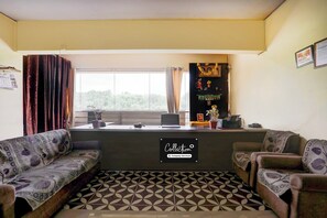 Um quarto com sofás estampados, uma escrivaninha com um computador, uma janela com cortinas e uma estante na parede com objetos decorativos.