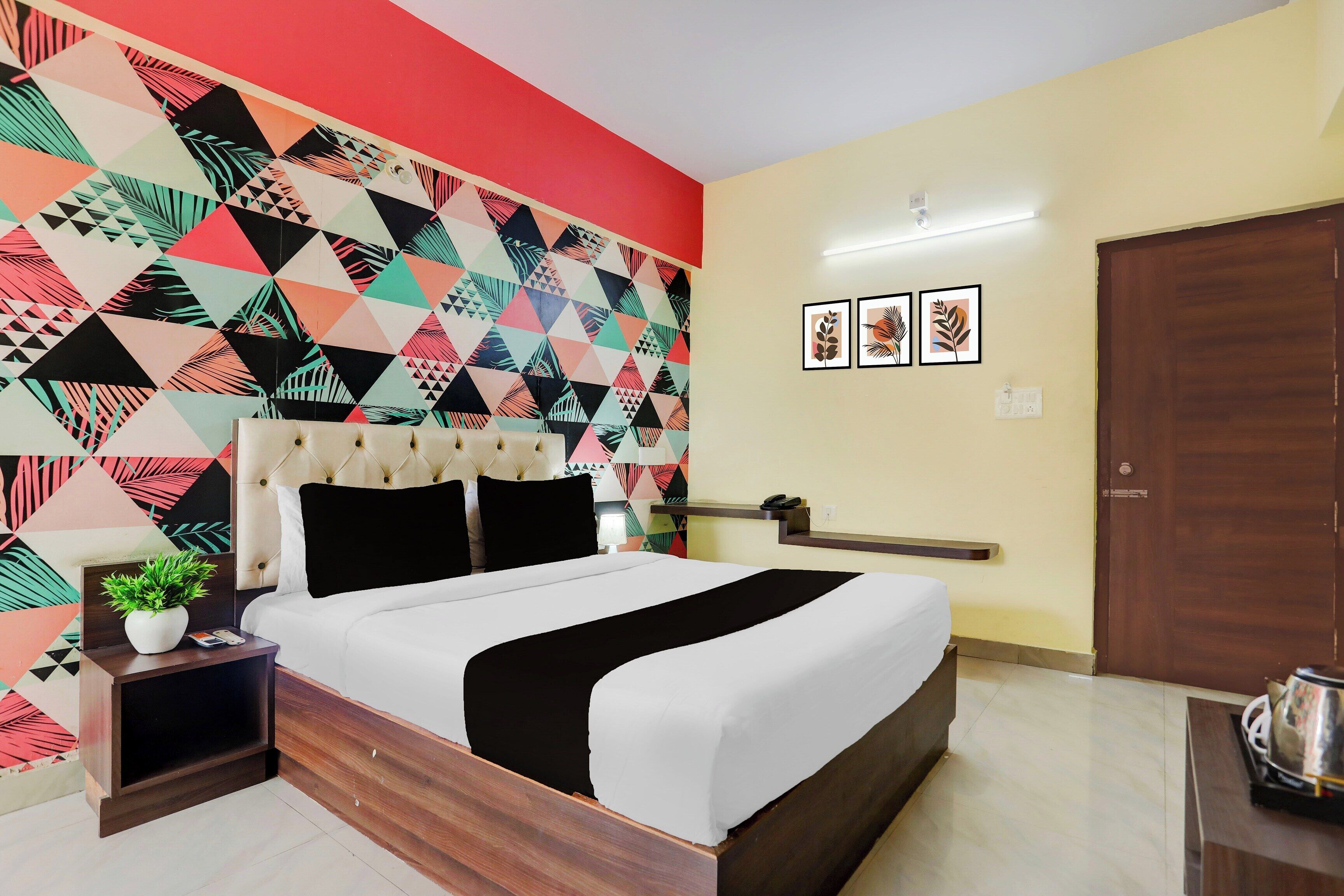 A hotel room with a bed, a nightstand, a plant, a wall with geometric patterns, and a door.