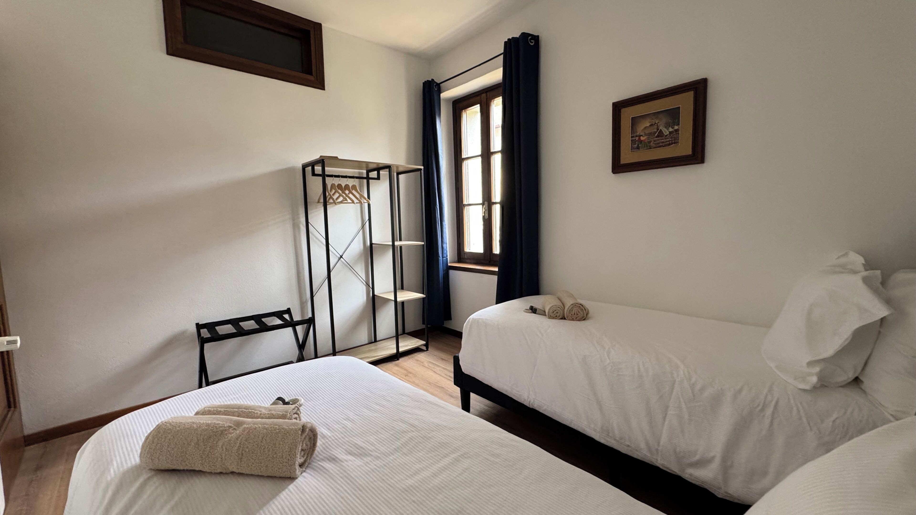 A bedroom with two beds, a window with curtains, a radiator, and a framed picture on the wall.