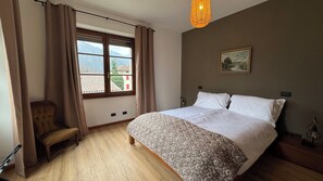 A bedroom with a large bed, a chair, a window with curtains, a painting on the wall, and a hanging light fixture. - La Casa del Re, just 400 meters from Lake Idro (Anfo)