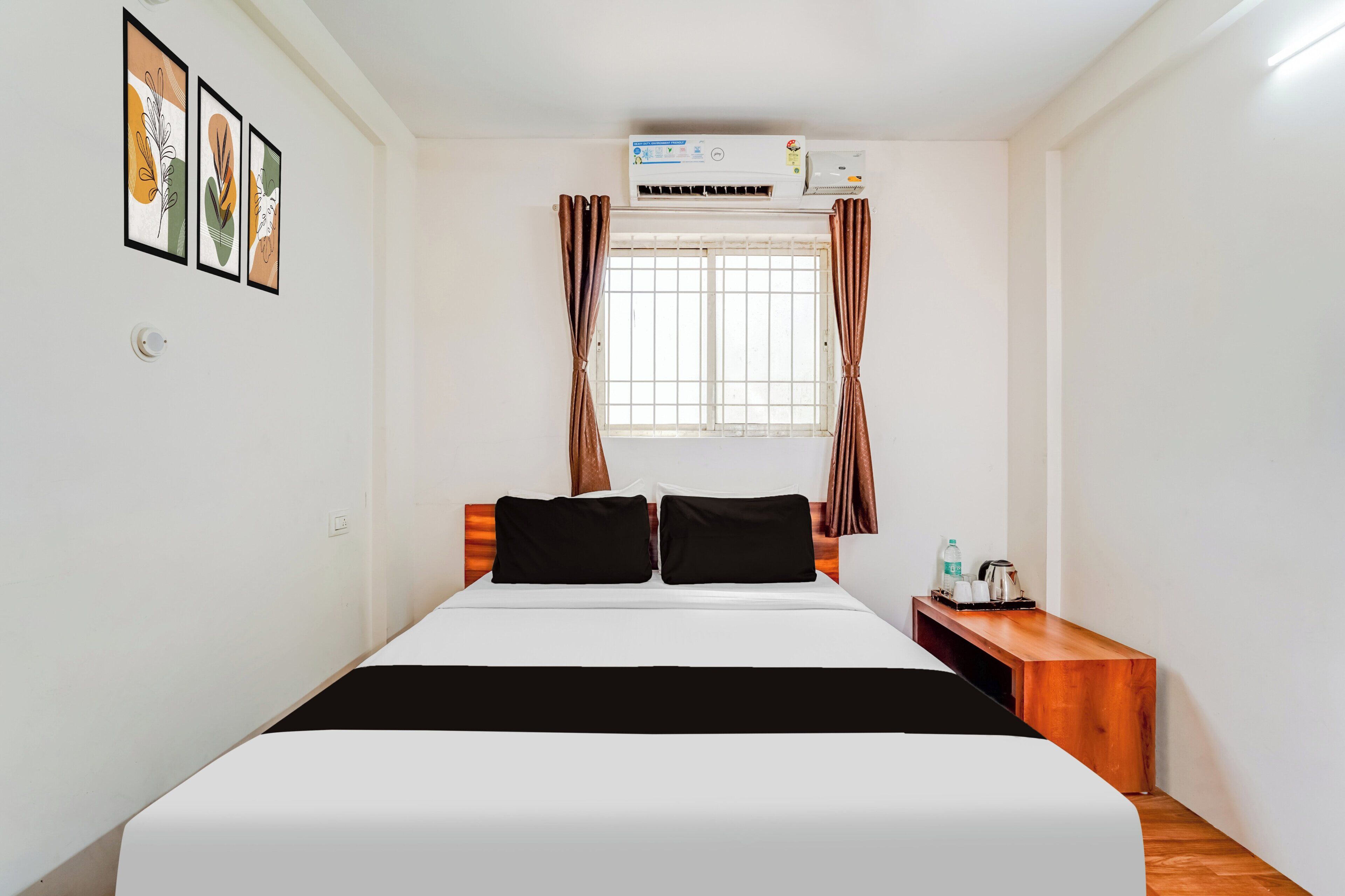 A hotel room with a bed, a window with curtains, an air conditioning unit, and a small wooden table with a kettle and cups.