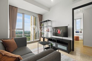 Ein modernes Wohnzimmer mit großem Fernseher, grauem Sofa und Blick auf die Stadt durch große Fenster.