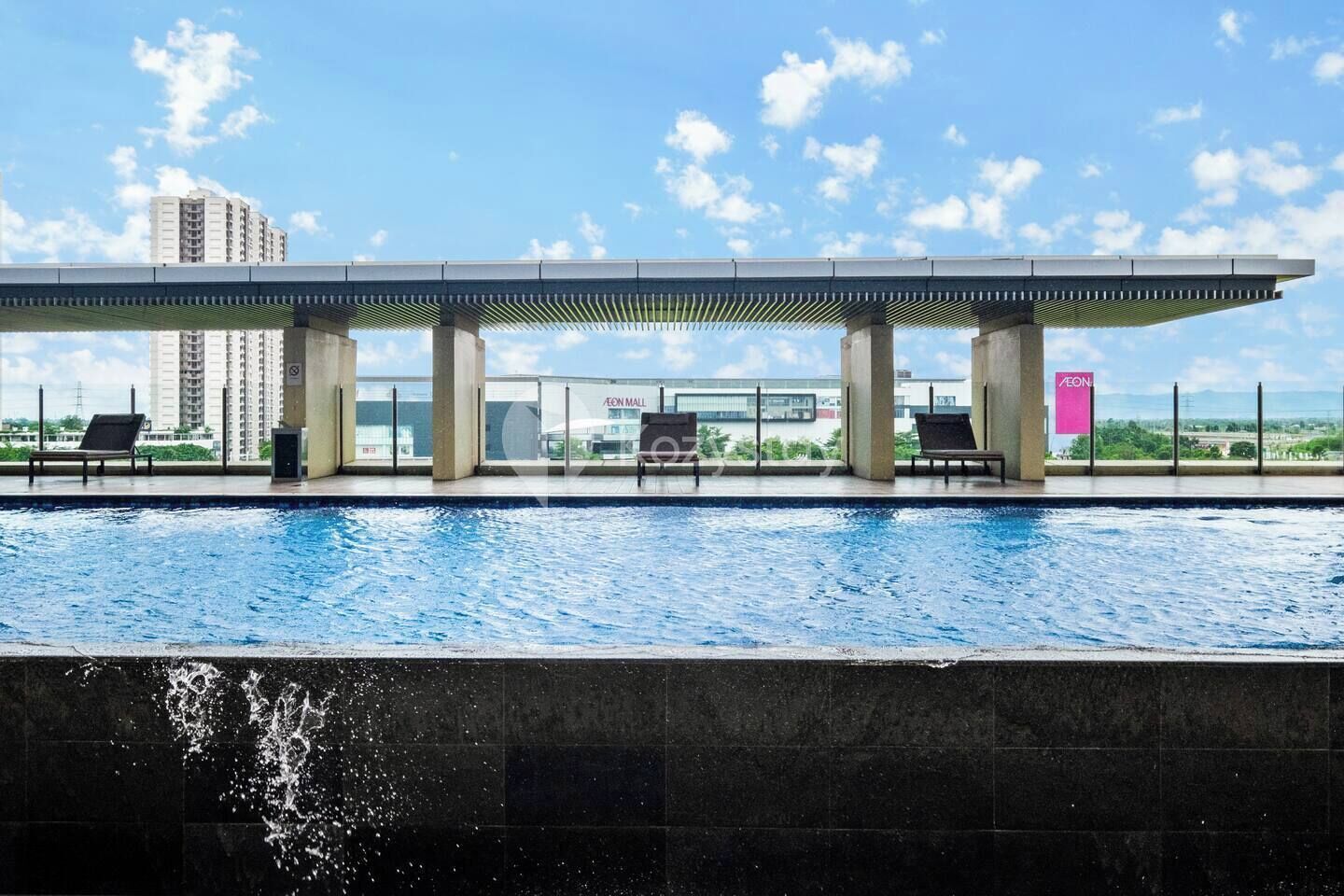 Ein Pool mit Liegestühlen, einem überdachten Bereich und Blick auf ein Einkaufszentrum sowie Gebäude im Hintergrund.
