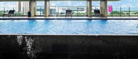 A swimming pool with lounge chairs, a covered area, and a view of a mall and buildings in the background.