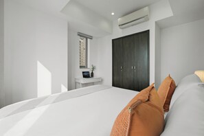 Ein modernes Schlafzimmer mit einem großen Bett, einem dunklen Wandschrank, einem kleinen Schreibtisch mit einem Laptop und einem Fenster mit Blick auf ein Gebäude.