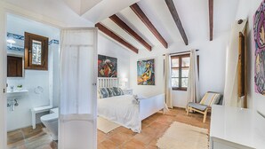 A bedroom with a bed, a bedside table, a window with curtains, a painting on the wall, and a bathroom with a sink and toilet. - Villa Near Pollensa Old Town (Pollença)