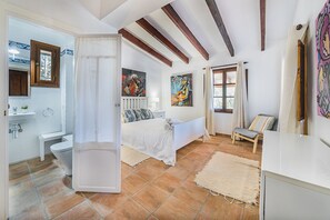 A bedroom with a bed, a bedside table, a window with curtains, a painting on the wall, and a bathroom with a sink and toilet. - Villa Near Pollensa Old Town (Pollença)