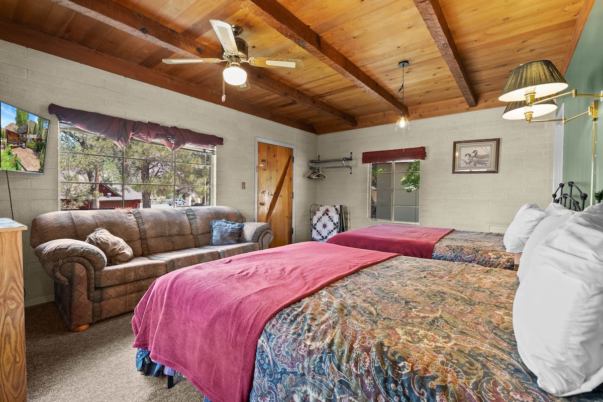 A room with a wooden ceiling, a ceiling fan, a bed with a patterned blanket, a couch with a red throw, and a window with a view of trees.