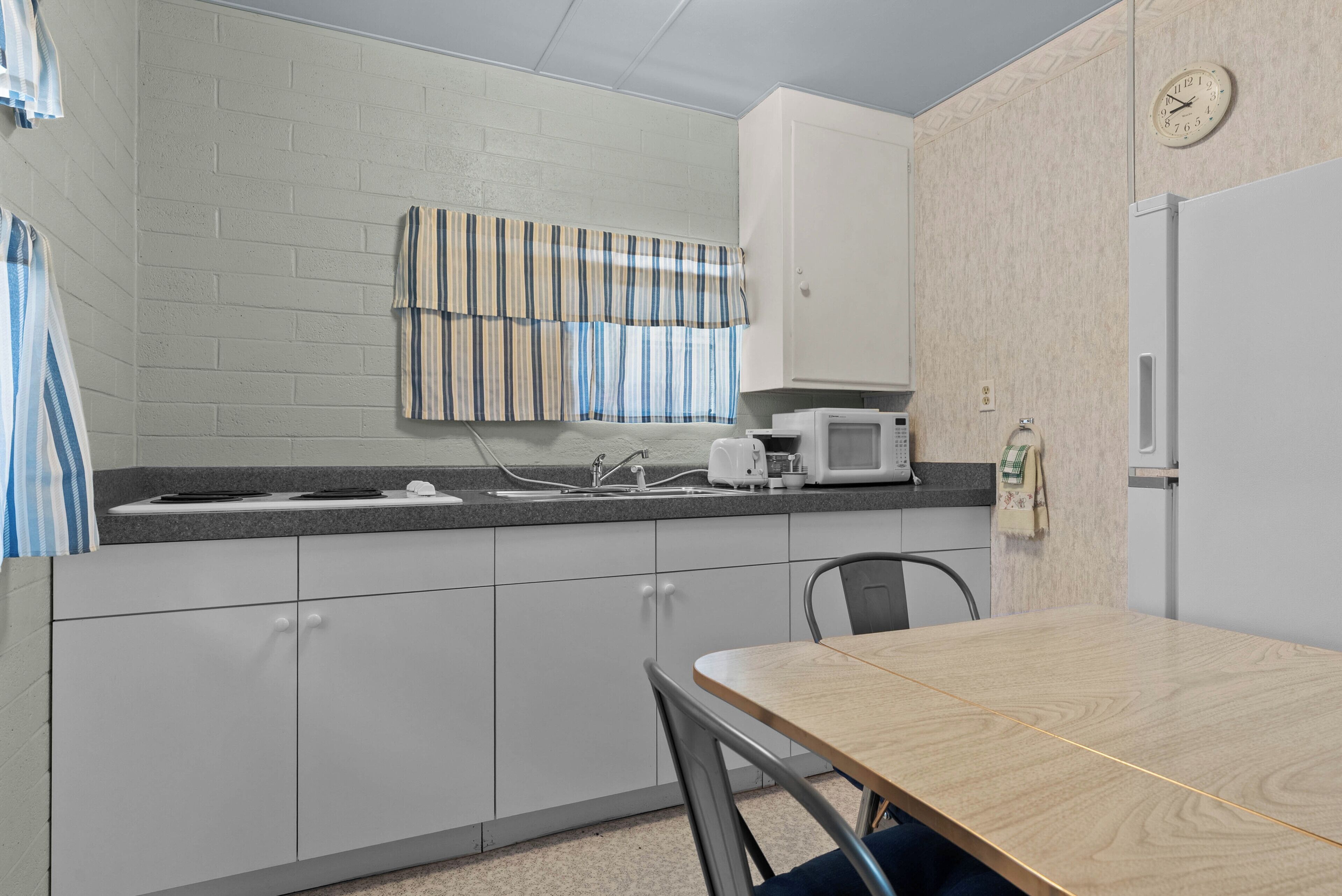 A kitchenette with a microwave, sink, and cabinets. There is a clock on the wall and a striped curtain over the window.