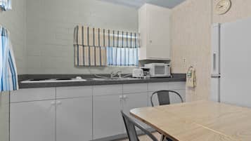 A kitchenette with a microwave, sink, and cabinets. There is a clock on the wall and a striped curtain over the window.