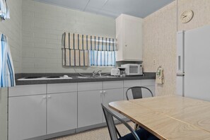 A kitchenette with a microwave, sink, and cabinets. There is a clock on the wall and a striped curtain over the window.