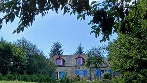 Un edificio de piedra con dos ventanas de tejado rojo y una chimenea, rodeado de vegetación.