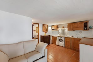 A compact kitchen with a washing machine, wooden cabinets, and a white tiled backsplash. - ‘Casa Vacanze Eucalipto’ Apartment with Sea View, Shared Pool, and Private Garden (Li Cuncheddi)