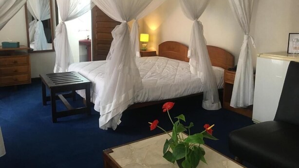 A bedroom with a four-poster bed, a bedside table, a nightstand, a chair, and a small table with a vase of flowers. - ivotel  (Antananarivo)