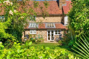 Ein Steinhäuschen mit Ziegeldach, ein Garten mit verschiedenen Pflanzen und eine Terrasse mit Gartenmöbeln.