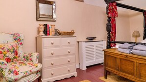 A bedroom with a floral-patterned armchair, a white dresser, a four-poster bed, and a mirror on the wall. - Finest Retreats | Lavender Cottage (Methwold)