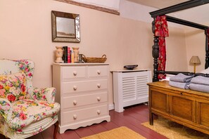 Ein Schlafzimmer mit einem floralen Sessel, einer weißen Kommode, einem Himmelbett und einem Spiegel an der Wand.
