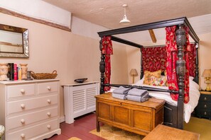A four-poster bed with red curtains, a wooden nightstand, a dresser with drawers, and a mirror on the wall. - Lavender Cottage (Methwold)