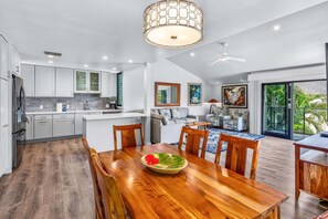 A modern kitchen with white cabinets, a dining area with a wooden table and chairs, and a living room with a sofa and a large window. - Wailea Elua 1104 - Ocean Views, Central A/C, Walk to Ulua Beach, Shops & Dining! (Kihei)